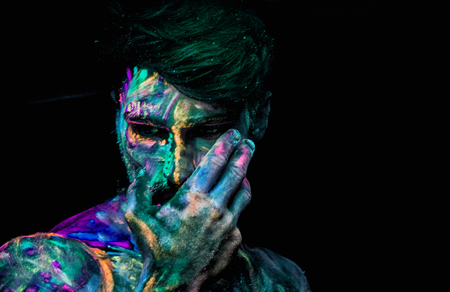 Headshot of young man painted in fluorescent paint on face and muscular torso, in studio shot with UV lightの写真素材