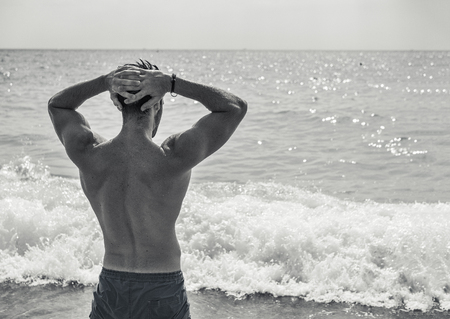 Handsome muscular young man standing on a beach, relaxed, taking a bath, wearing bathing suit, seen from the backの写真素材