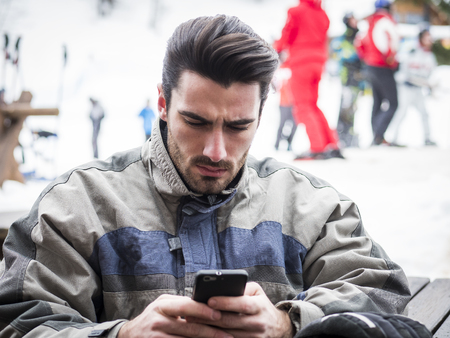 Handsome young man in skiing suit relaxing, typing text message or surfing the internet, sitting at table outside ski resortの写真素材
