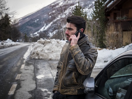 Smiling man talking on cellphone with thumb up near car with open hood. Snowy forest on backgroundの写真素材