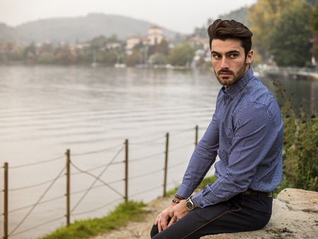 Portrait of contemplative handsome brown haired young man, with short beard, wearing shirt beside picturesque river or lake, looking away to a side during fallの写真素材