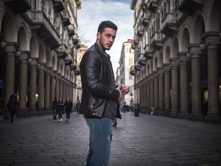 One handsome young man in urban setting in old classic European city, standing, wearing black leather jacket and jeans, looking at camera in Turin, Italyの写真素材