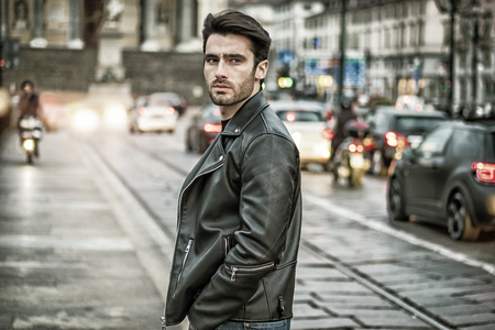 One handsome young man in urban setting in modern city, standing, wearing black leather jacket and jeans, looking at cameraの写真素材