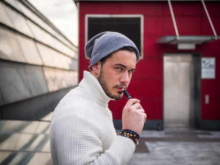 Young man vaping, smoking e-cigarette outdoor, wearing wool sweater and beanie cap