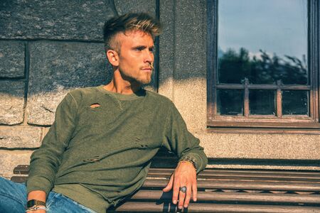 Handsome blond, blue eyed young man on bench outdoor and looking to a sideの写真素材