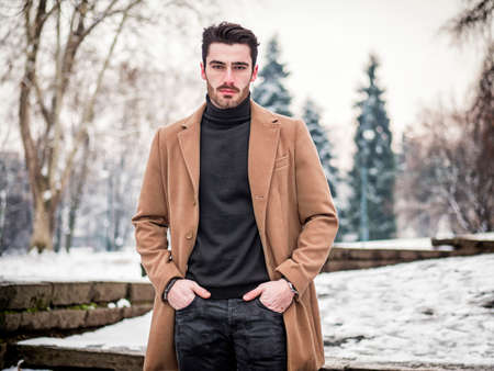 Young man in snowy city park wearing wool coat, looking at cameraの写真素材