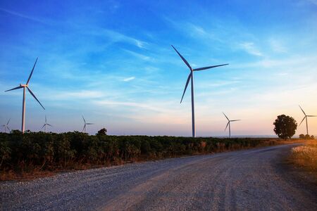 Rural road in a field with wind turbines at the sky.