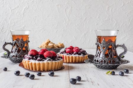 OTea cups, tarts cakes sweets with fresh raspberries and blueberry on white wooden background.