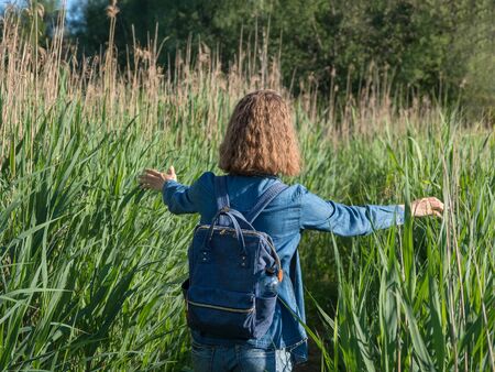 A view from the back of a blonde girl in a denim dress with a blue  backpack on her back, walking along a path in the tall grass. Travelling in wild.の写真素材
