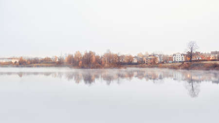 Panorama of a misty morning minimalistic landscape with a line of cottages and trees by the water in autumnの写真素材