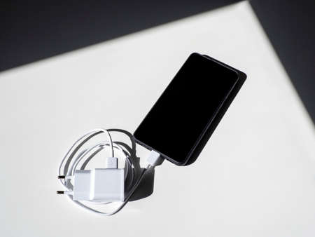 A black smartphone with a charging wire on a white table with shadows. Top view.