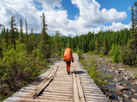 A male tourist with a large orange backpack walks along an old wooden bridge against the background of a coniferous forest and mountains in the distanceの写真素材