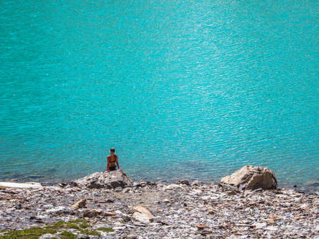 Back view girl in bikini. Sexy tanned girl in sits on lake rocks. Female in bathing suit sits on...