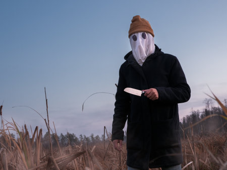 Faceless killer. Dangerous man with a white cloth on his face and a knife in his hand on the reeds in the evening. Halloween.の写真素材