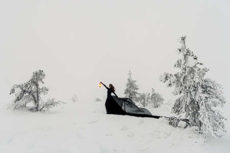 Fairy beautiful woman in a long black dress holds an old lantern in her hand in a snowy forest against the background of a foggy winter hill and snowfall. Black witch comes out of the snowy forest.の写真素材