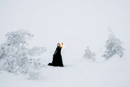 soft focus. Young beautiful woman in long black dress with old lantern over winter hill background and snowfall. Fairy tale girl on polar winter landscape. Black witch in the snow.の写真素材