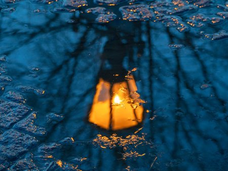 Blurred antique lantern shines yellow and is reflected in a spring puddle. Urban postcard abstract view.の写真素材