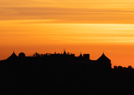 Contrasting view of dark silhouette of the roofs against the bright orange evening sky. Low angle view of long row of characteristic townhouses with roofs and chimneys of Europe houses during sunset.の写真素材