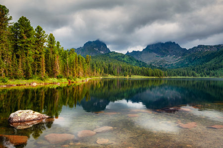 Picturesque shore of a mountain lake with cedars under a dramatic sky. Taiga rocks with blue sky in the warm sun and trees Mountain sunny landscape. Ergaki Nature Park in the mountains of Siberia.の写真素材