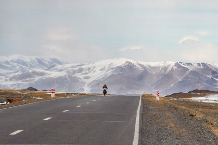 Mongolian motorcyclist in the distance on a mountain highway. Snow straight winter road through the hills. Mountain road on a cloudy winter day.の写真素材
