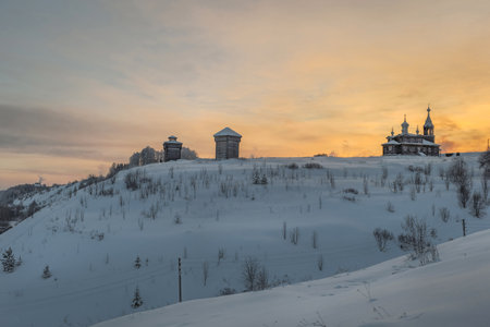 St. Elijah's Church and Trinity hill in Cherdyn. Ancient architecture is surrounded by tall white drifts and orange sunset skies. Historical center of the ancient city of Cherdyn, Russia of winter.の写真素材