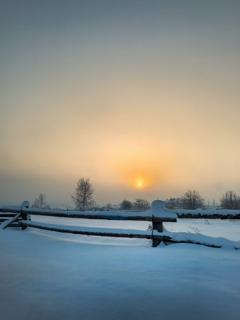 Beautiful winter morning. Frosty sunrise over the village. Misty sunrise over the field with wooden fence and village in the background. Vertical view.の写真素材