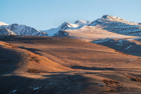 Scenic view to snowy mountain ridge illuminated by setting sun under clear sky in golden sunset tones. Evening alpine landscape in sunset color. Light and shadow in mountains. Perfect image for wallの写真素材
