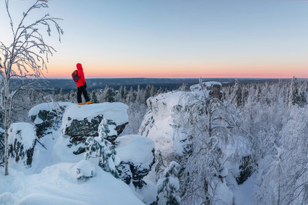 Mystical dawn in the snow-covered mountains. Man tourist stands over the winter cliff and looks into the distance. High stone cliff covered with snow and rise above the coniferous forest at sunset.の写真素材