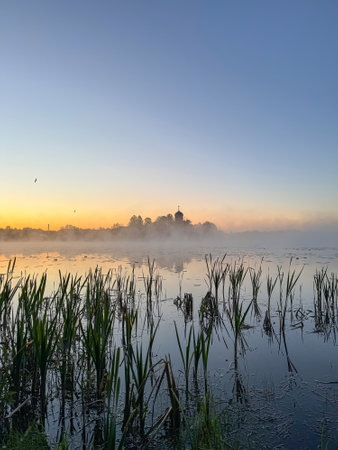 Morning landscape, sunrise on the river. Wide river, horizon, clouds are reflected in the water. Vertical view of the monastery in foggy morning. Golden Ring of Russia.の写真素材
