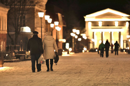 Selective focus. Age couple, unrecognizable view from the back, walking in the snow between the main square and the main street of Rybinsk, during a winter snowy night in Yaroslavl region. Russia.の写真素材