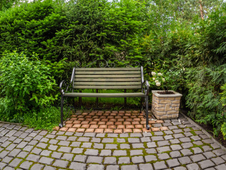 A wrought-iron bench in the shade of large bushes at the back of the garden. Flowers in a pot in a garden in sunlight in summer.の写真素材