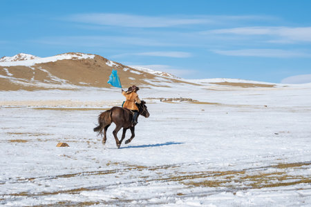 Unidentified young boy with a flag in his hand ride a horse across the snowy steppe. An...