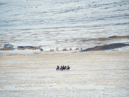 Mongolian horsemen on the background of yurts and snow-capped mountains. Difficult living conditions, windy steppe, harsh nature. View from distance.の写真素材
