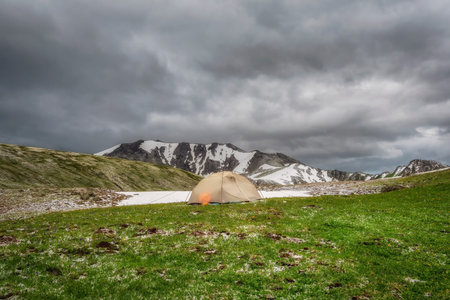 Awesome mountain scenery with tent. Terektinsky ridge, Altai. Green glade high in the mountains is covered with hail. Dark stormy sky. Solo hike.の写真素材