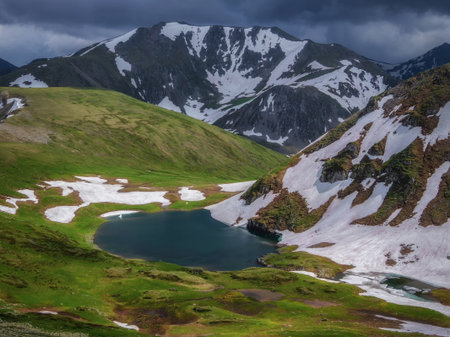 Mountain lake in highlands. Beautiful small lake among green grassy hills and rocks with view to snow-capped mountain range. Scenic landscape with alpine lake and snowy high mountains.の写真素材