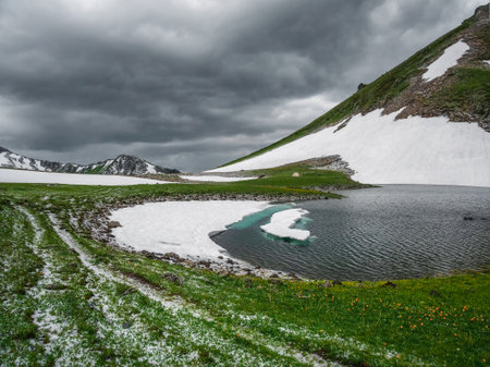Mountain lake in highlands. Beautiful small lake among green grassy hills and rocks with view to snow-capped mountain range. Scenic landscape with alpine lake and snowy high mountains.の写真素材