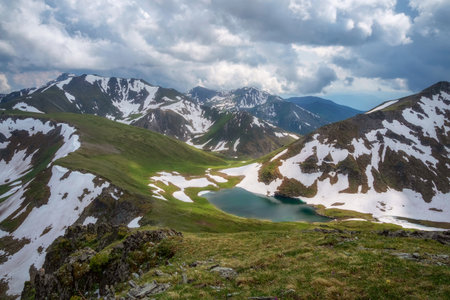 Amazing small lake among green grassy hills and rocks with view to snow-capped mountain range....
