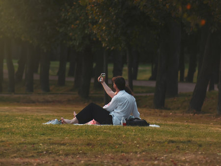 Soft focus. A couple sits on the grass in a summer park in the evening and takes a selfie.の写真素材