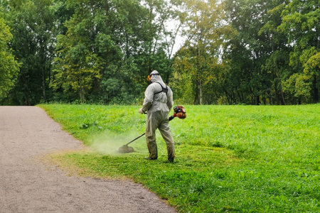 Unrecognizable man in uniform mows the lawn in city park. Mowing the grass with a lawn mower. Garden...