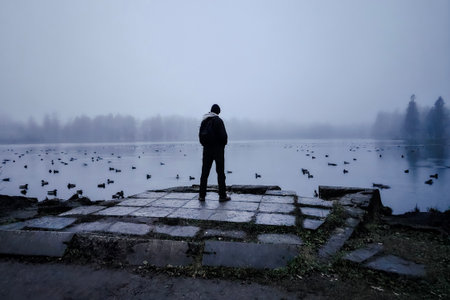 The tall man looks into the distance, gloomy photo. Misty blue winter landscape with lake and islands in fog. An unrecognizable view from the back.の写真素材