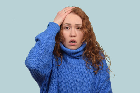 Portrait of young redhead woman with shocked facial expression, keeps hands on head, dressed in blue...