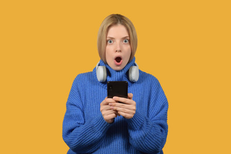 Surprised young woman in earphones holding smartphone and looking at camera isolated over yellow backgroundの写真素材