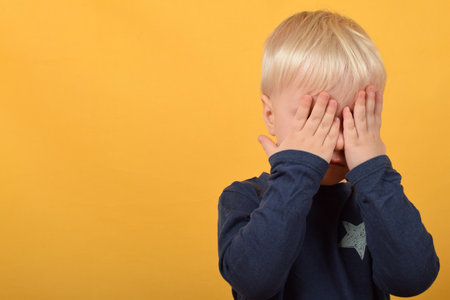 Little boy covering his face with his hands, isolated on yellow background