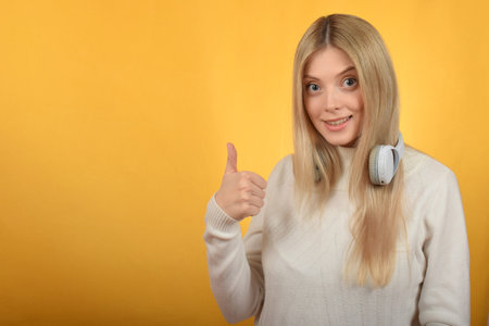 Portrait of beautiful young woman with headphones showing thumbs up on yellow backgroundの写真素材