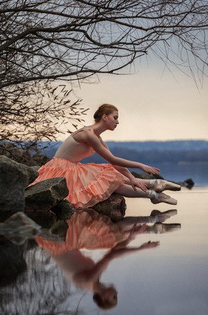 Ballerina in ballet pose sits above the lake on background of sky, trees and large stones. Her reflection can be seen In water.の写真素材