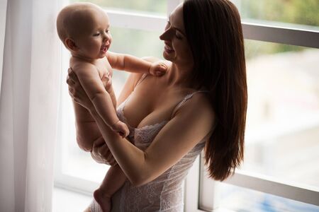 Young mother playing with her baby. They are sitting near window and laugh gaily.の写真素材