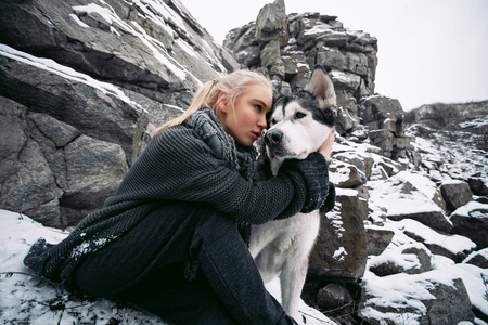 Girl with dog Malamute among rocks in winter. They sit on rocks among scattering of stones and snow. Girl hugs a dog's neck.の写真素材