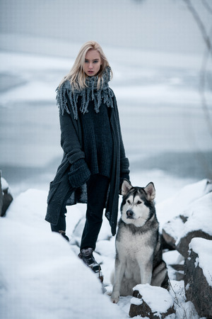 Girl with dog Malamute among rocks in winter. She stands on snow among rocks. Dog sits next to girl.の写真素材