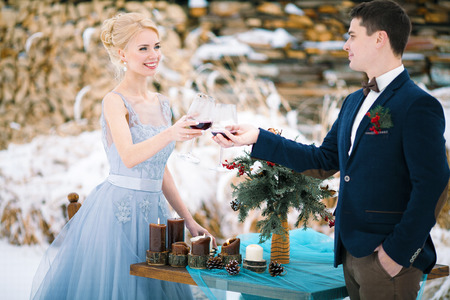Winter wedding outdoor on table and firewood background. Bride and groom stand near table and clink glasses with wine. Beside them is table with bouquet.の写真素材