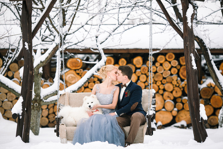 Bride and groom in winter with dog sitting on swing and a kiss. Dog sitting next to them.の写真素材